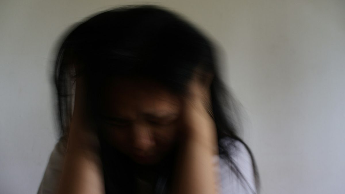 Mulher levando as mãos à cabeça em imagem com leve desfoque, contexto de sintomas de migrânea